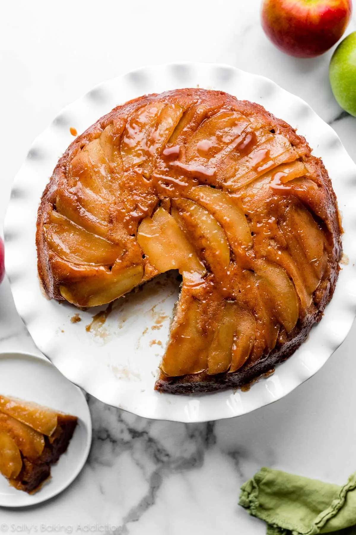 Golden apple upside-down cake with a caramel glaze, served on a white pie plate with a slice removed.